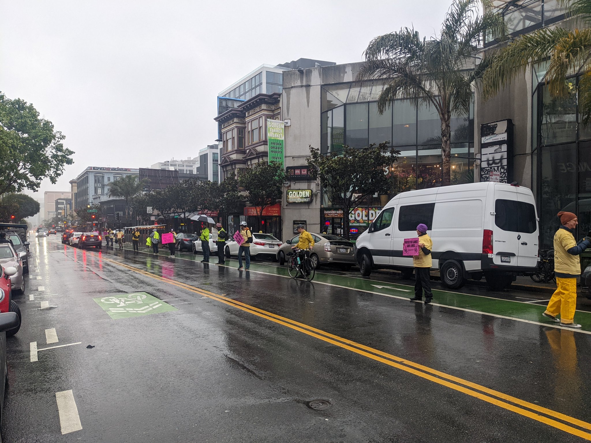 A dozen people, mostly in yellow shirts, stand between the green bike lane on Polk street and the road in the rain, keeping cars out of the bike lane.