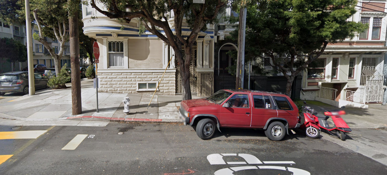 A photo of a street. A narrow strip of pavement is painted red, and a small yellow strip on a stop sign indicates it's a bus stop. A car is parked in front of the bus stop.