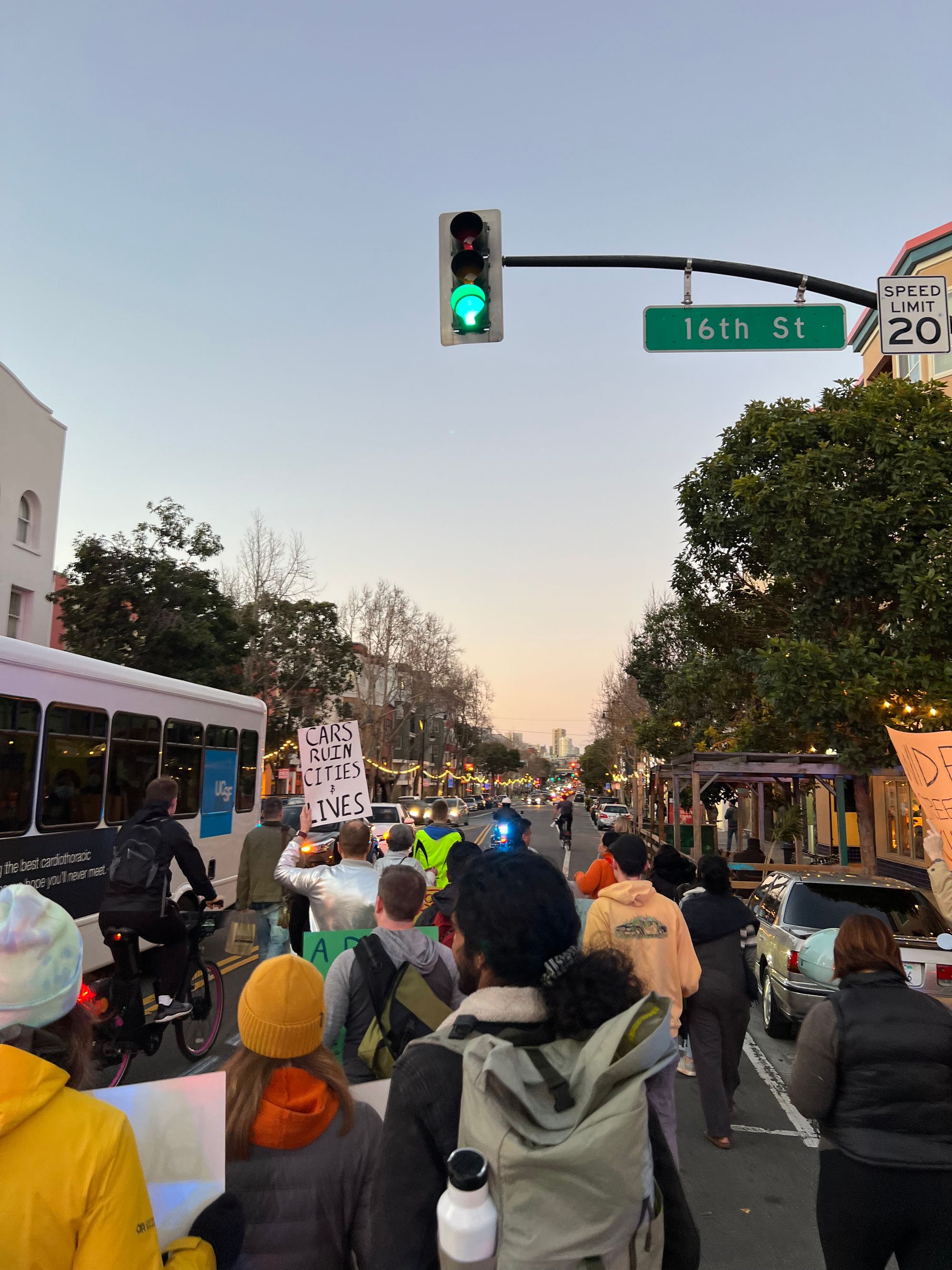 a group of people marching and biking down 16th street in the Mission. One person is holding a sign that says, “Cars Ruin Lives and Cities”