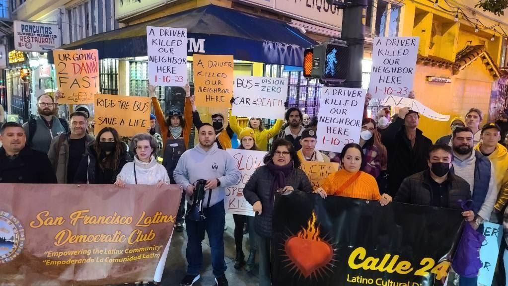 A group of people at a street corner holding signs “U R going 2 Damn fast” “ride the bus, save a life” “ a driver killed our neighbor here 1-10-23” “bus = vision, zero car = death” “Calle 24” “San Francisco Latinx Democratic Club”
