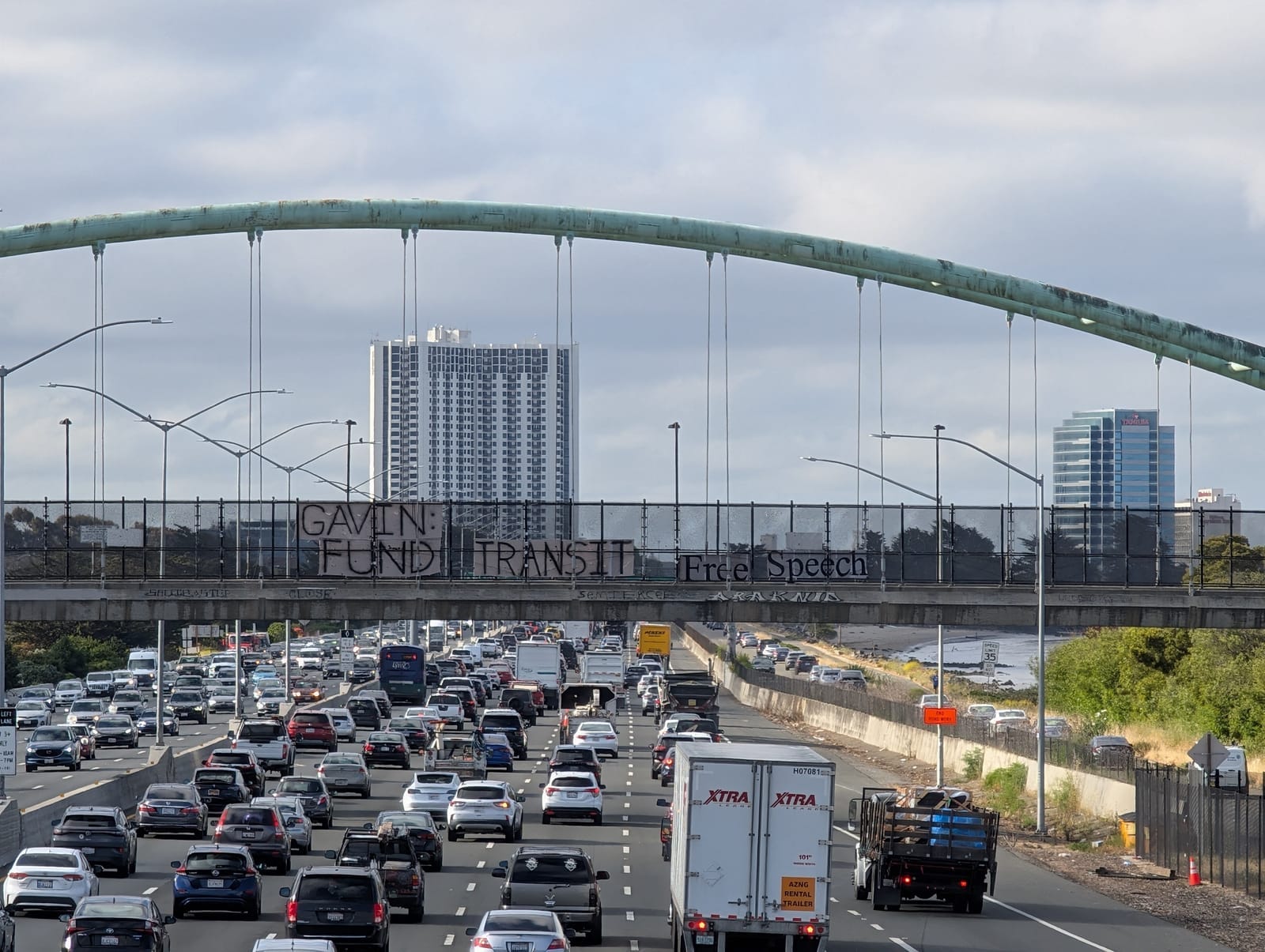 Banner above a traffic-ridden freeway in Berkeley draped on a pedestrian bridge reading "Gavin: Fund Transit"