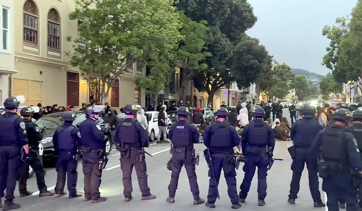 dozens of police form a large circle around what looks like a dozen teenagers who are sitting down in the street