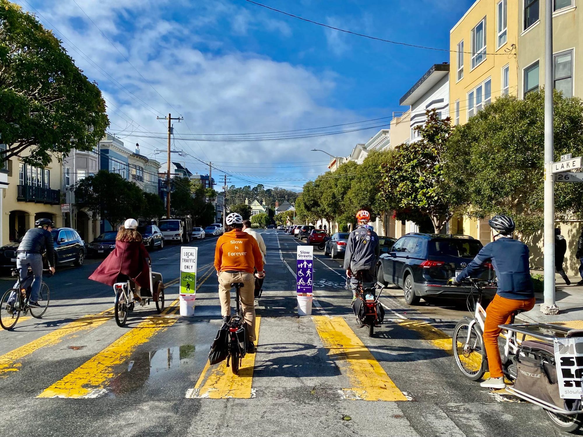 A group of bikers pass by unofficial Slow Street signs in buckets filled with concrete. The signs resemble SFMTA Slow Street signs. The bikers are riding cargo bikes.