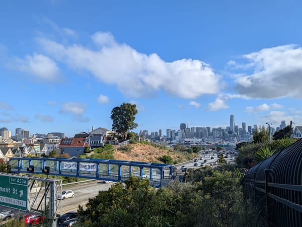view of a banner reading "Gavin: Fund Transit 4ever" on a pedestrian walkway over the highway with a view of downtown San Francisco in the background